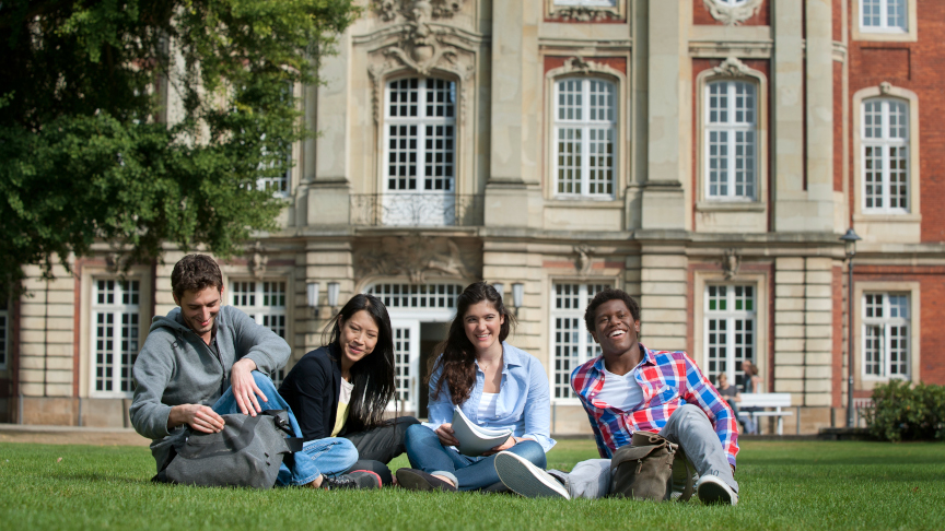 Four students are sitting on a meadow in front of a university